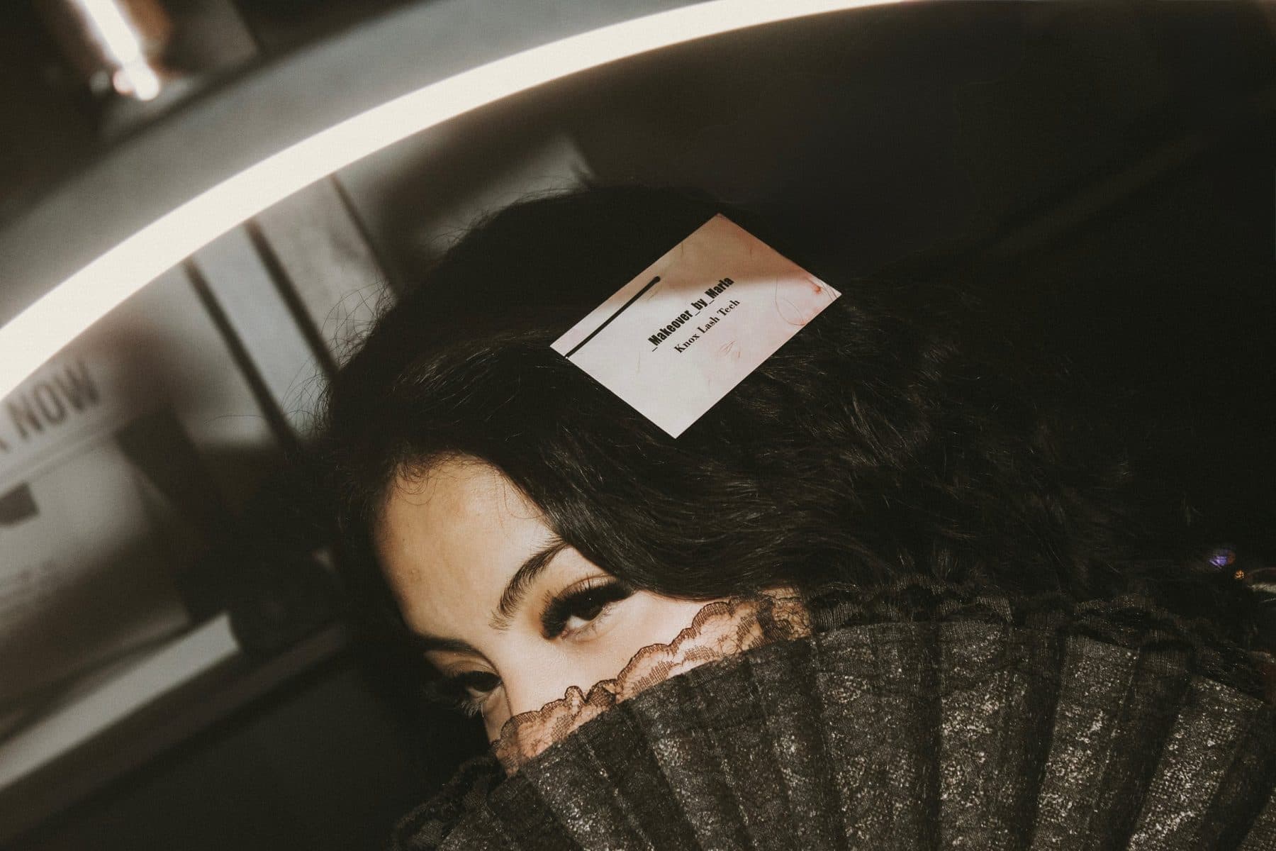 Maria looking over a black lace fan, her Makeover by Maria business card tucked in her hair, lash extensions visible